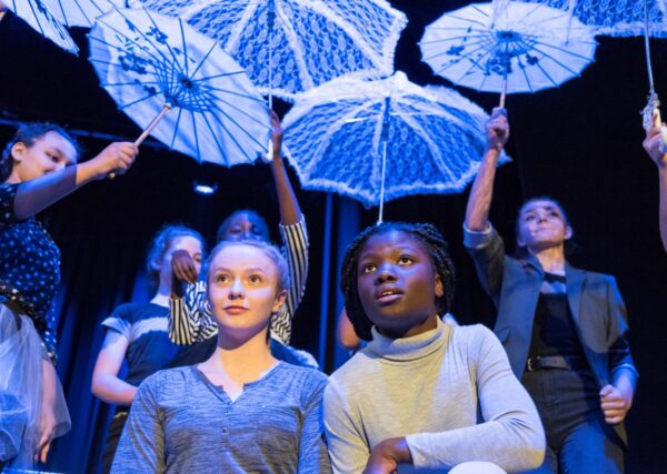 A group of children in a CSSF drama performance hold up umbrellas in the background. Two girls sit at the front in the centre of the picture looking up. One is black with dark hair and one is white with blonde hair.