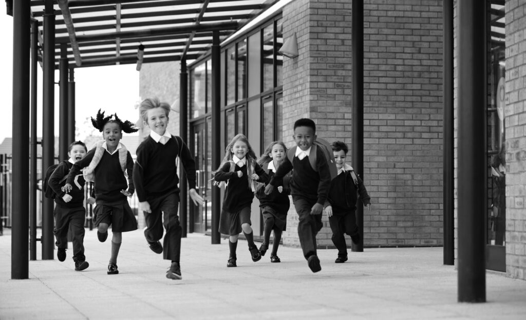 Primary school kids, wearing school uniforms and backpacks, running on a walkway outside their school building, front view